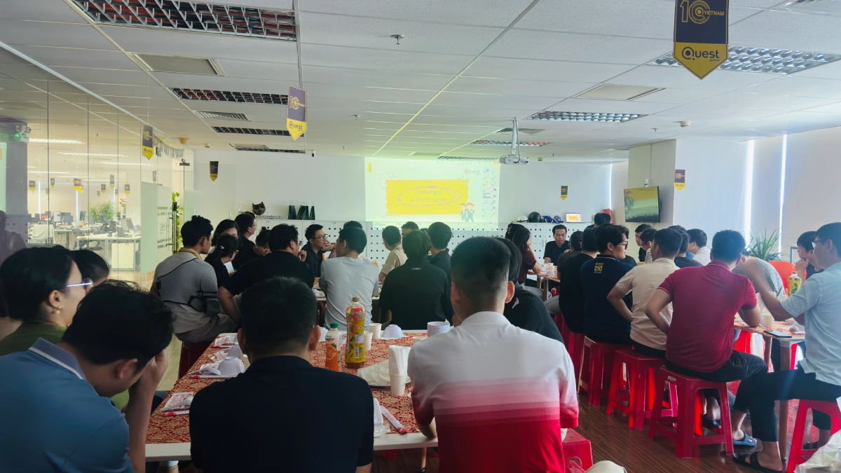 Employees gathered at long tables for an office social event or casual meeting, viewing a presentation projected onto a screen